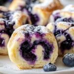Golden blueberry cheesecake rolls with glaze and powdered sugar on a wooden plate