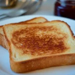 Two slices of golden-brown toast on a white plate with jam and a knife nearby.