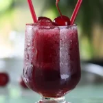 A Cherry Rum and Coke Slushy in a tall glass, garnished with cherries and a lime wedge, with a blurred summer patio background.