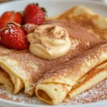 A stack of rolled Cinnamon Roll Protein Crepes drizzled with cream cheese glaze on a rustic wooden table.