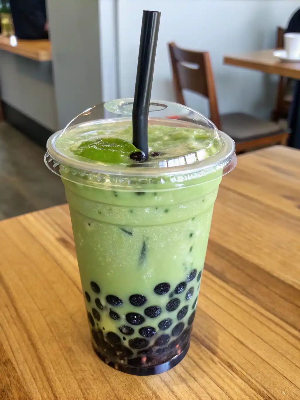 Matcha Green Tea Boba Ingredients Iced matcha green tea boba in a clear plastic cup with black tapioca pearls and a paper straw, surrounded by matcha powder and ingredients.